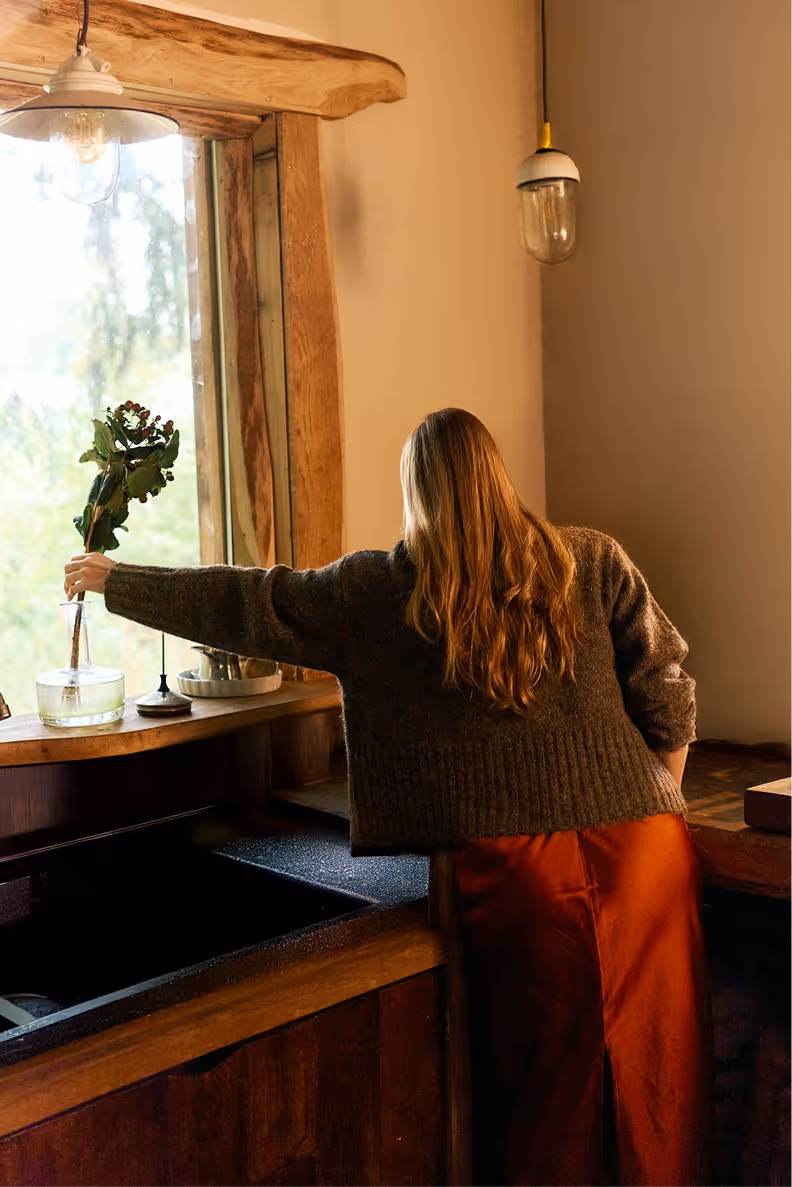 Woman with long hair in a cozy sweater placing a vase with green leaves and berries on a wooden windowsill in a rustic kitchen.