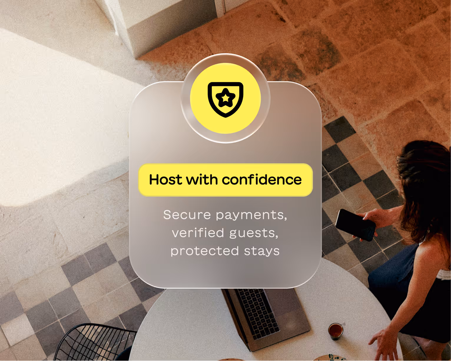 Top view of a woman holding a phone near a round table with a laptop and coffee, with a text overlay saying Host with confidence. Secure payments, verified guests, protected stays.