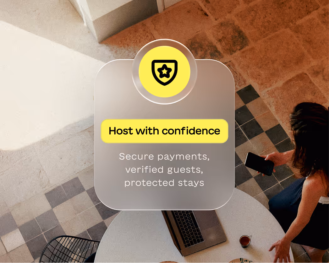 Overhead view of a person holding a smartphone next to a table with a laptop and coffee, with a highlighted text box reading 'Host with confidence, Secure payments, verified guests, protected stays.'