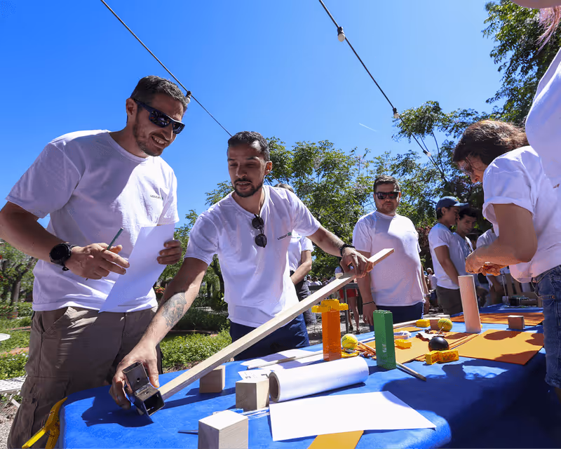 Group of people outdoors at a table with wooden blocks and papers, collaborating on a hands-on project under clear blue sky.