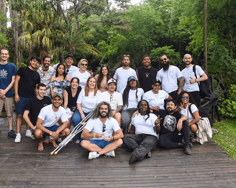 Group photo of twenty diverse people smiling outdoors on a wooden deck surrounded by green trees and plants.