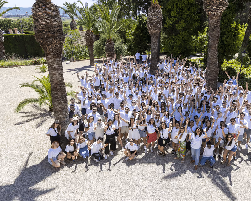 Large group of people wearing white shirts posing and waving outdoors among palm trees on a sunny day.