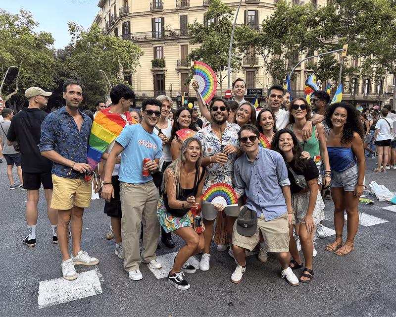 Group of cheerful young people posing on a city street during a pride event, some holding rainbow fans and a rainbow flag.