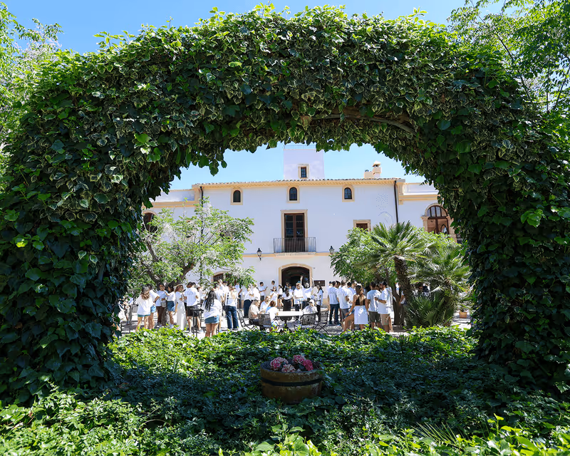 Group of people gathered outdoors in front of a white building framed by a large leafy green arch.