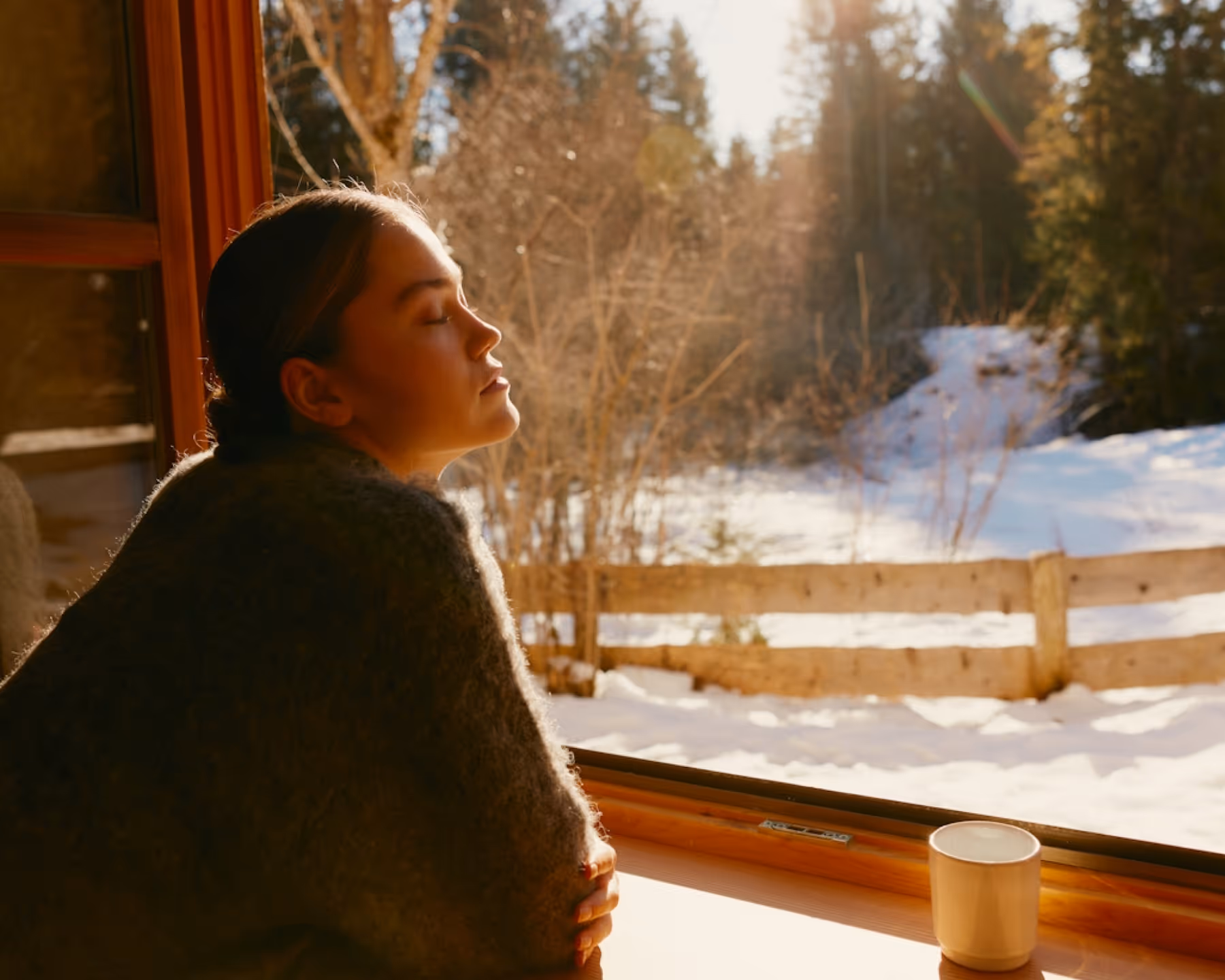 Woman with closed eyes enjoying sunlight by a window overlooking a snowy yard with wooden fence and trees.