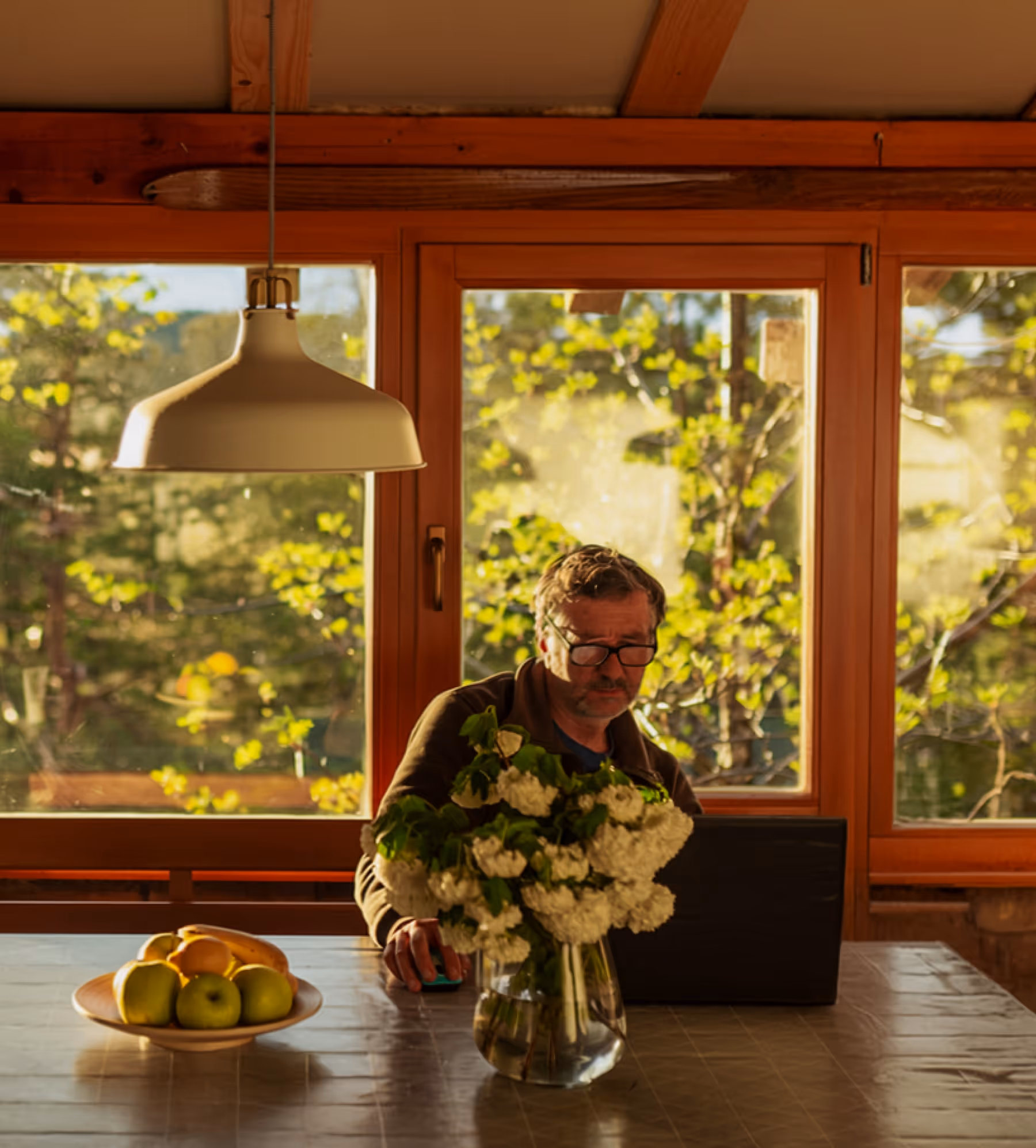 Man wearing glasses working on a laptop at a table with a vase of white flowers and a bowl of fruit near a large window.