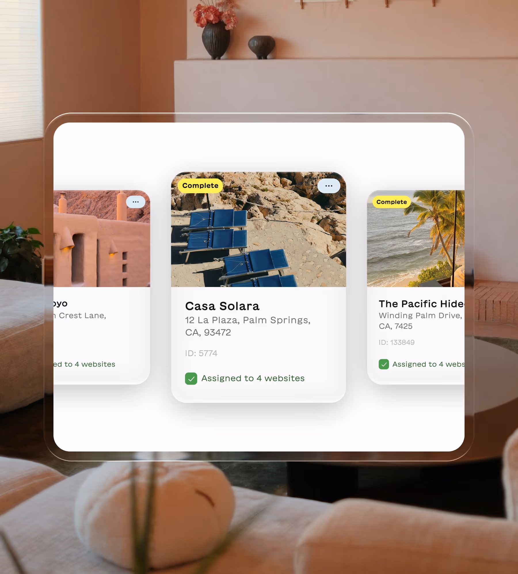 Three property cards displayed on a transparent screen in a cozy room, featuring Casa Solara in Palm Springs with blue lounge chairs on rocky terrain, each marked complete and assigned to 4 websites.