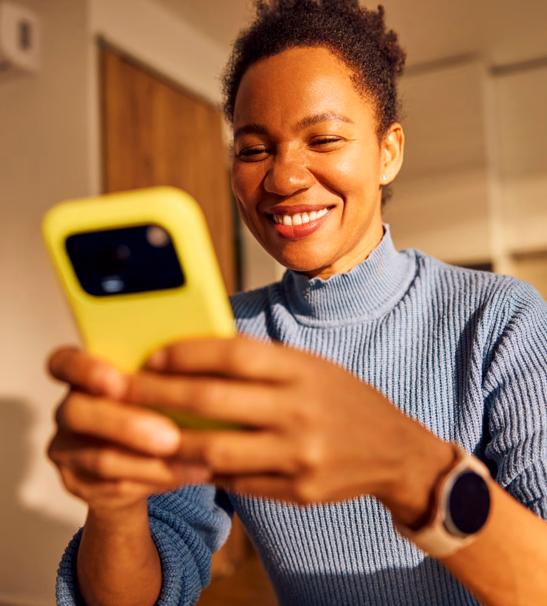 Smiling woman in a blue sweater looking at a yellow smartphone.