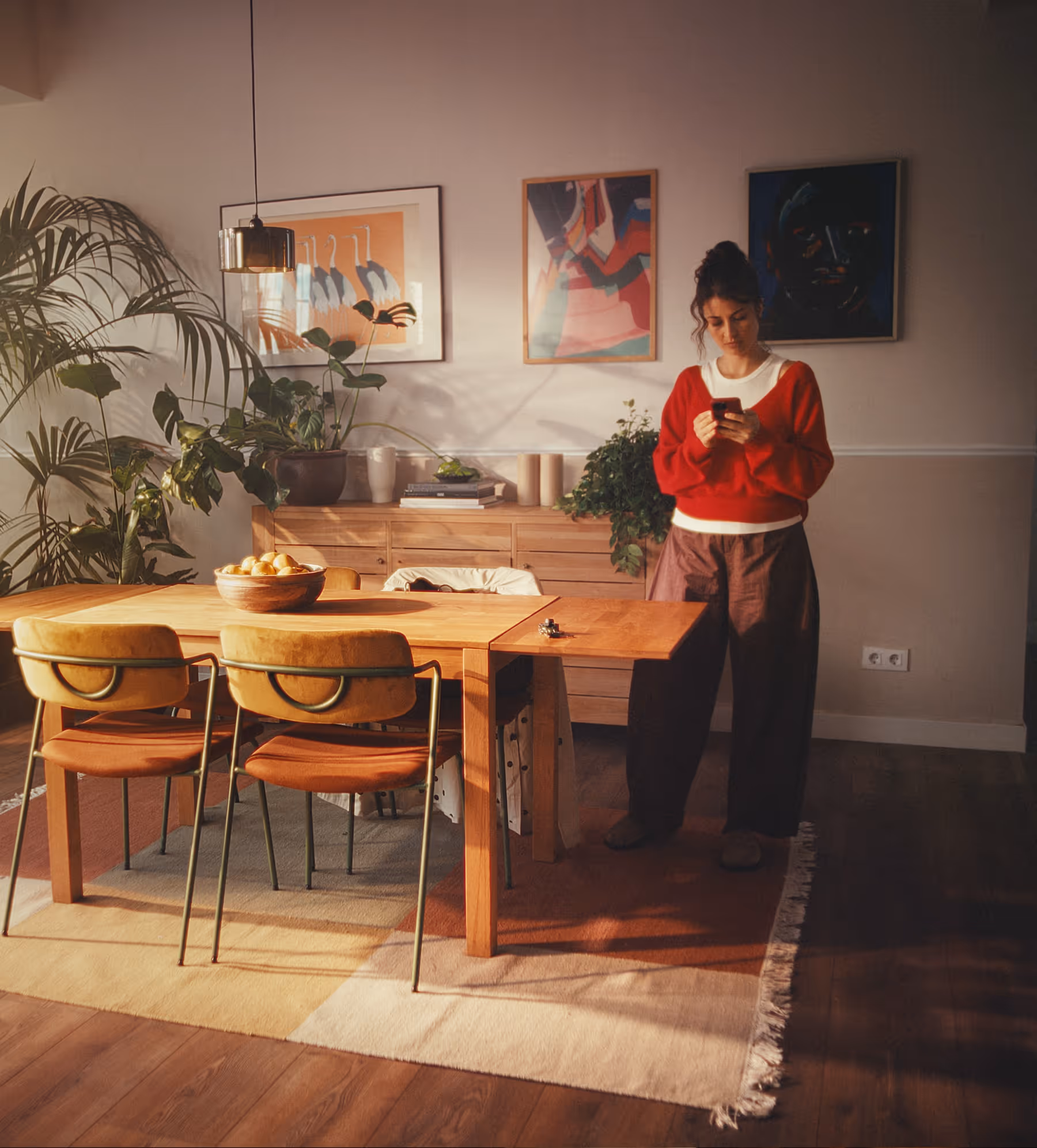 Woman in red sweater standing in a sunlit dining room with wooden table, chairs, indoor plants, and colorful wall art.