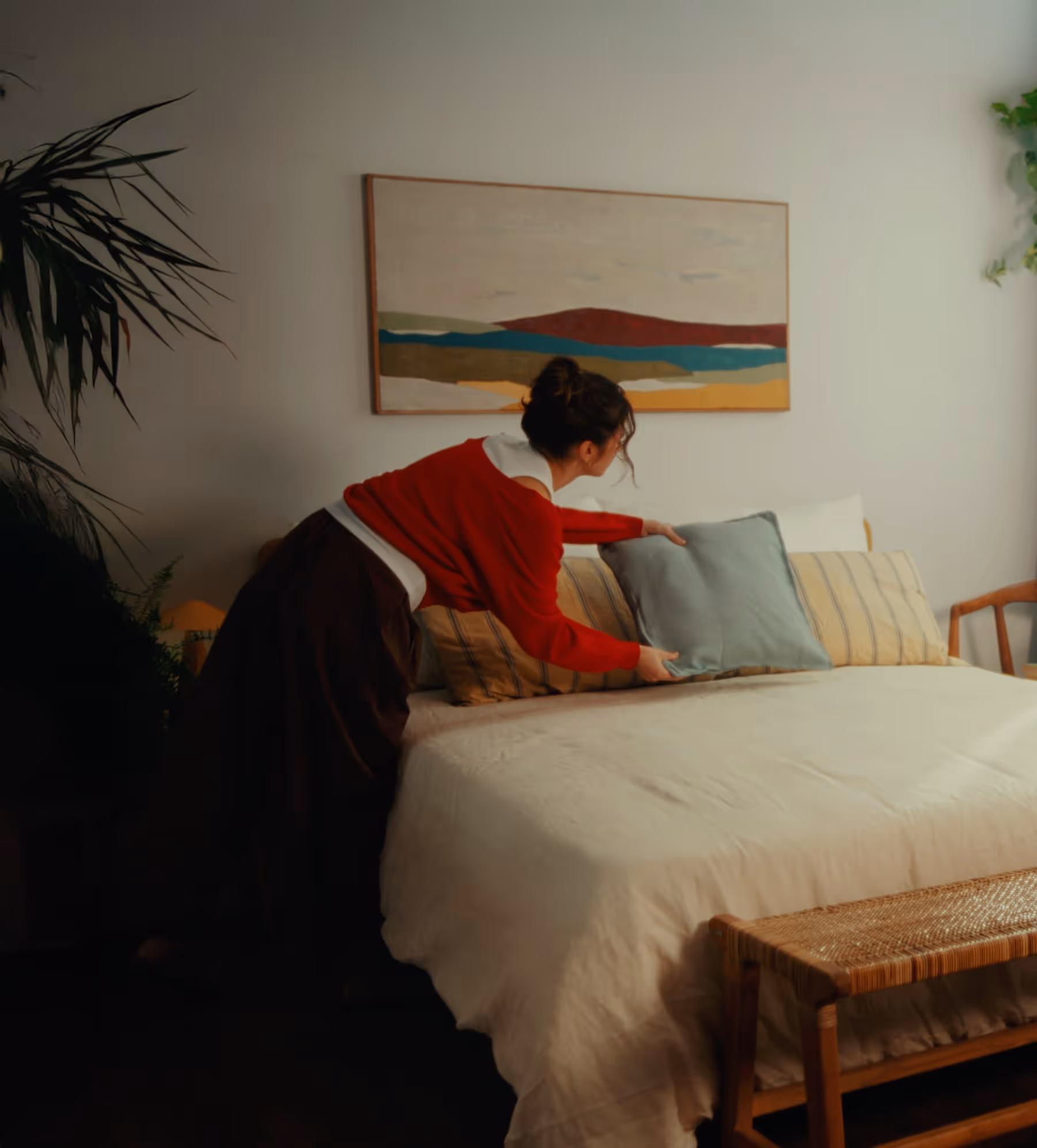 Woman in a red sweater arranging pillows on a neatly made bed in a cozy bedroom.