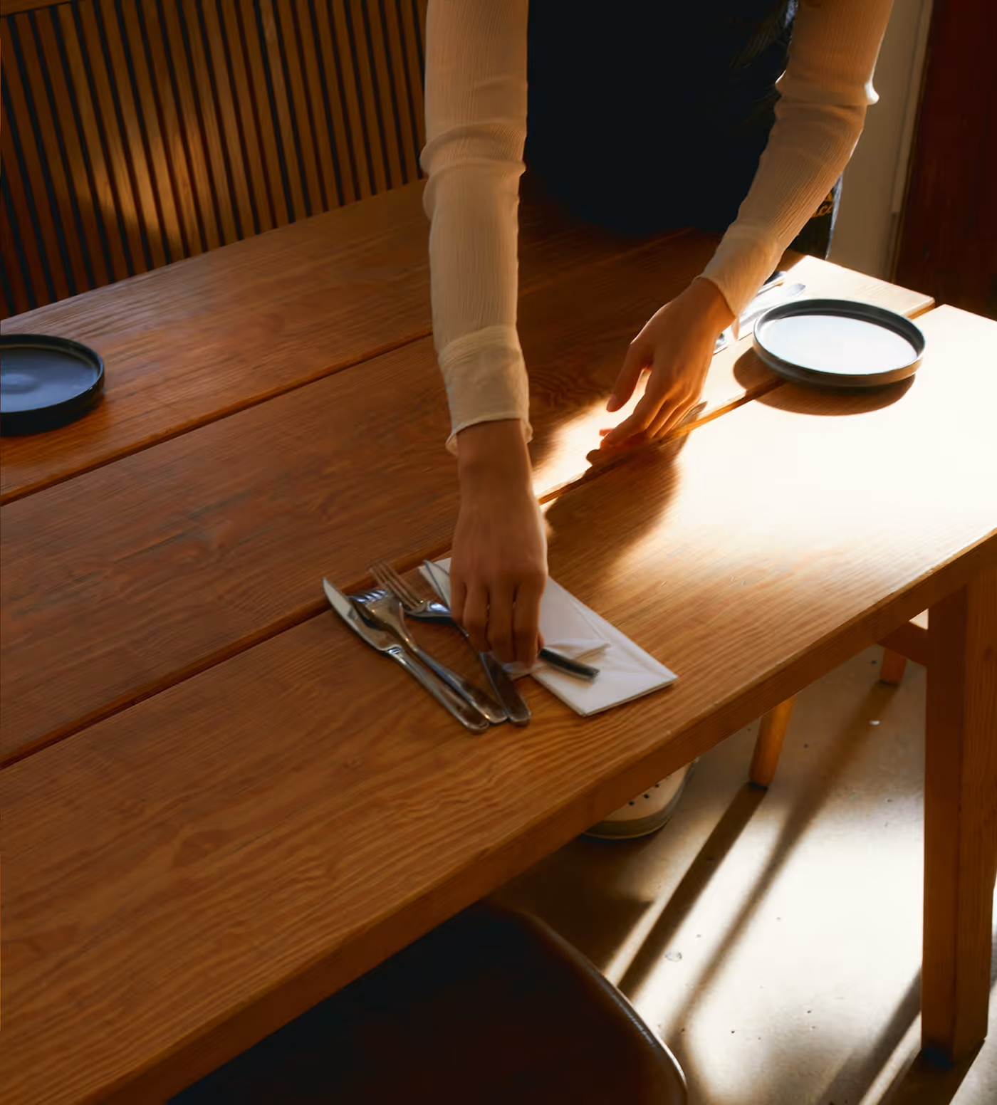 Person setting a wooden table with silverware and plates in warm natural light.