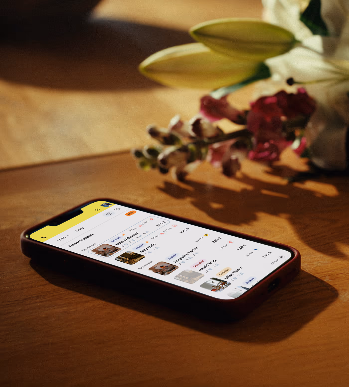 Smartphone on wooden table displaying a guest management app with reservations list and photos.