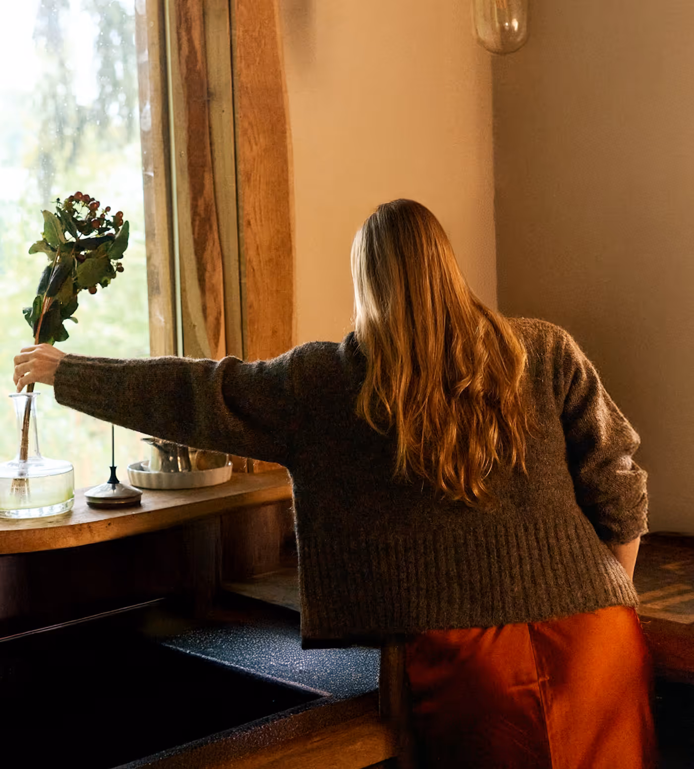 Person with long hair and brown sweater placing a bouquet with berries into a glass vase by a window.