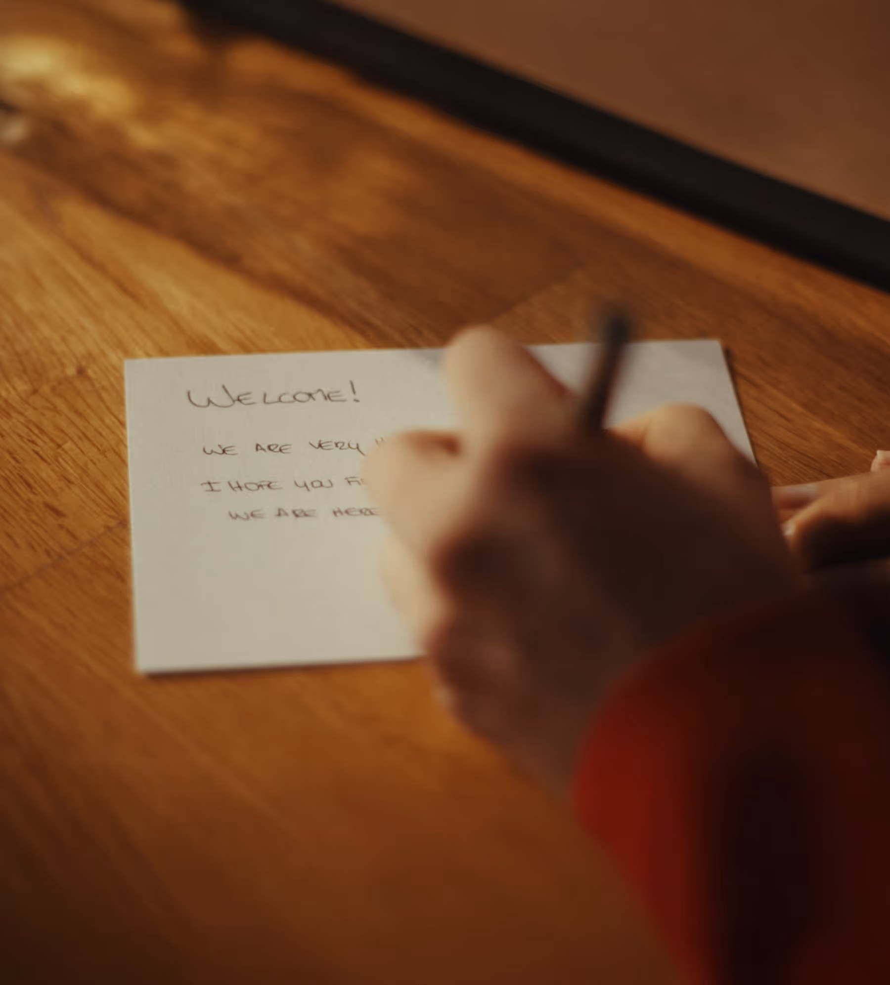Person writing a welcome note on a small card placed on a wooden surface.