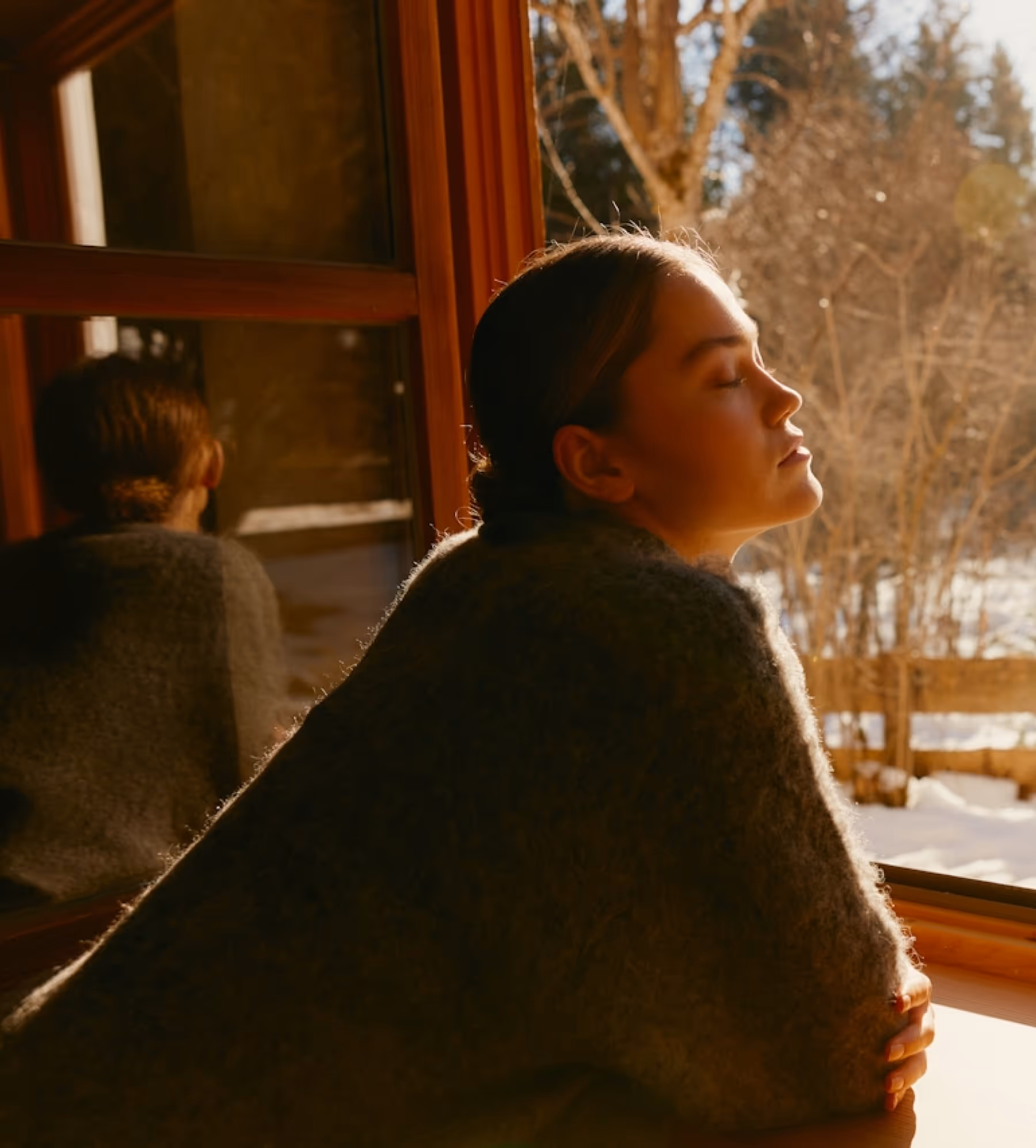 Woman with closed eyes basking in warm sunlight by a window with snow and trees outside.