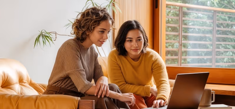 Two women sitting on a sofa looking at a laptop together in a cozy, well-lit room with a large window.