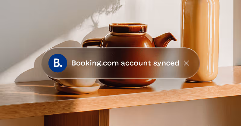 Notification popup on a wooden shelf with brown teapot, white cup on saucer, and beige vase saying 'Booking.com account synced.'