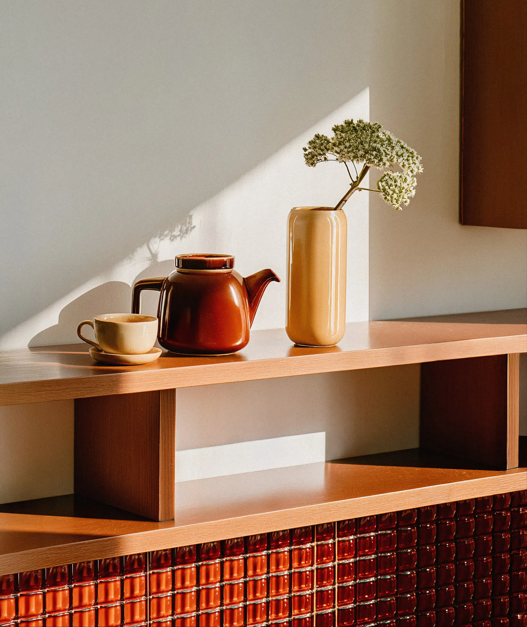 Brown ceramic teapot, beige cup with saucer, and tall beige vase with white flowers on a wooden shelf with light casting shadows.