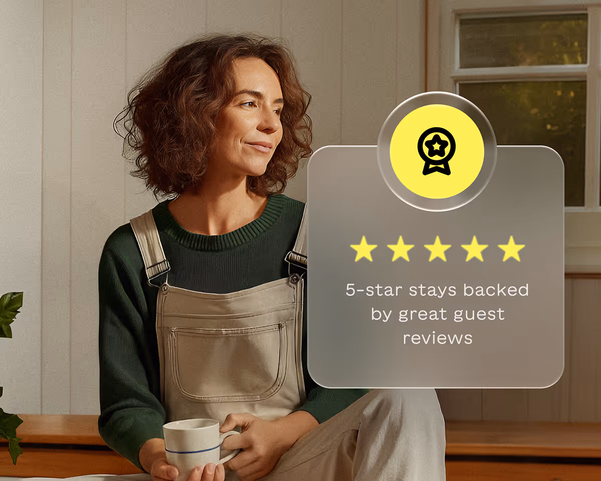 Woman with curly hair in beige overalls and green sweater holding a mug, sitting indoors beside a window and five yellow stars with text reading '5-star stays backed by great guest reviews.'