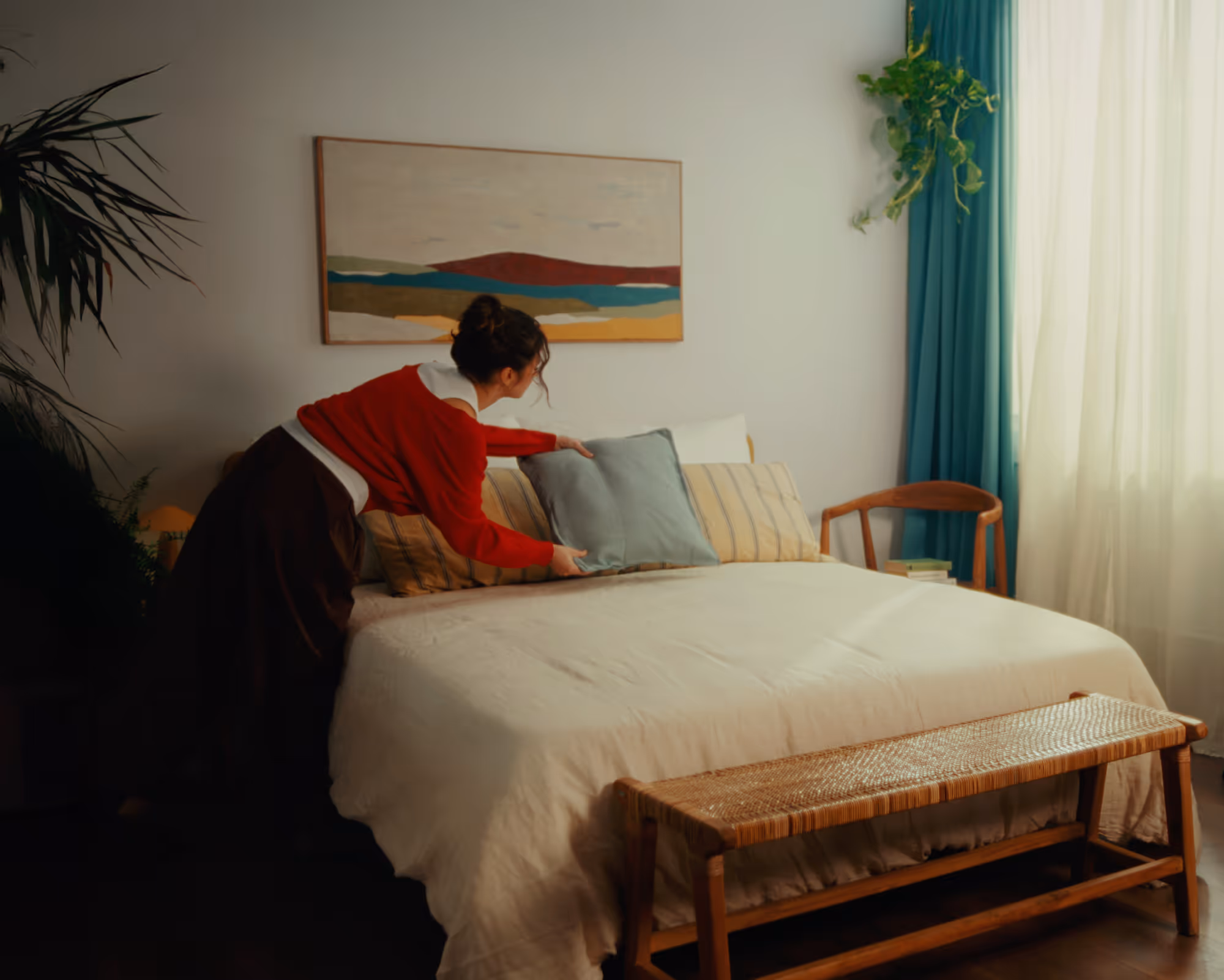 Person arranging a pillow on a neatly made bed in a cozy bedroom with indoor plants and a wooden bench.
