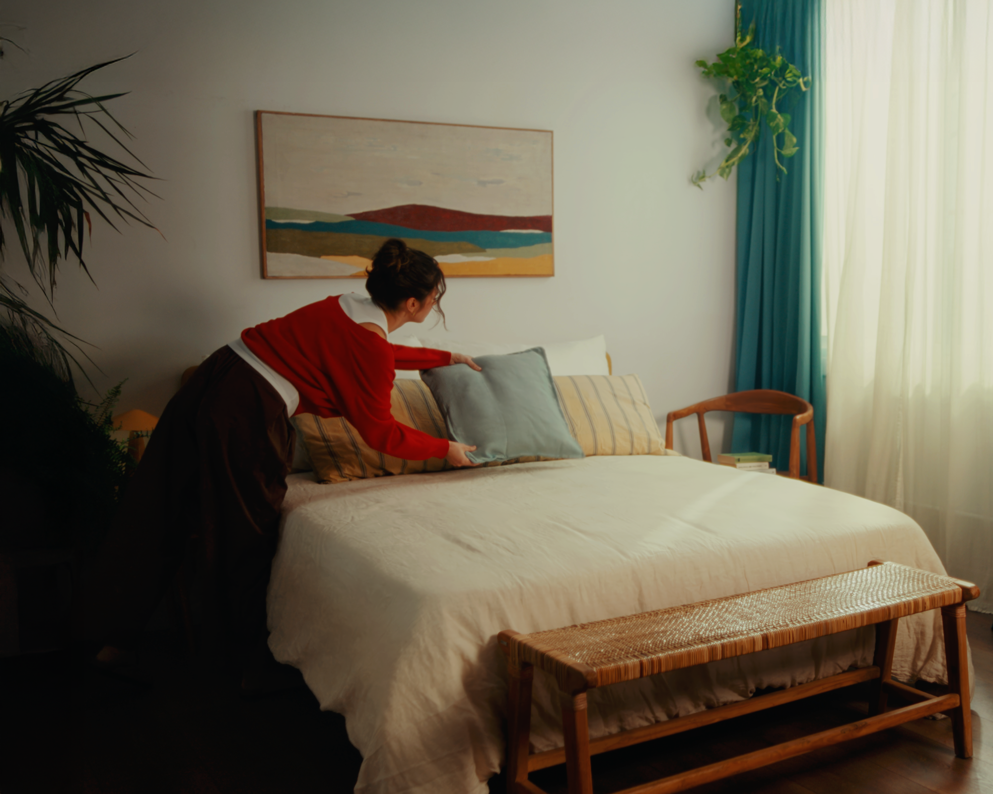 Person arranging a pillow on a neatly made bed in a cozy bedroom with indoor plants and a wooden bench.
