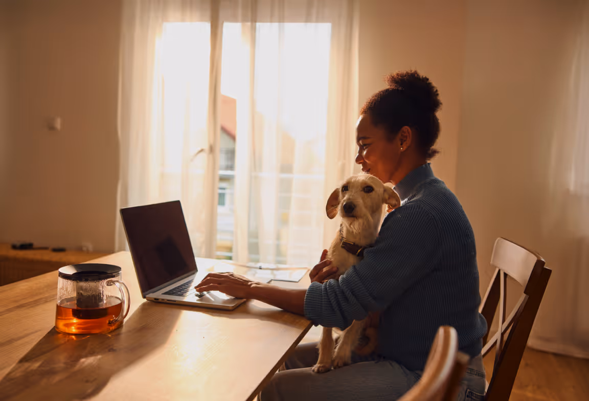 Woman sitting at a wooden table using a laptop while holding a small dog in a sunlit room.