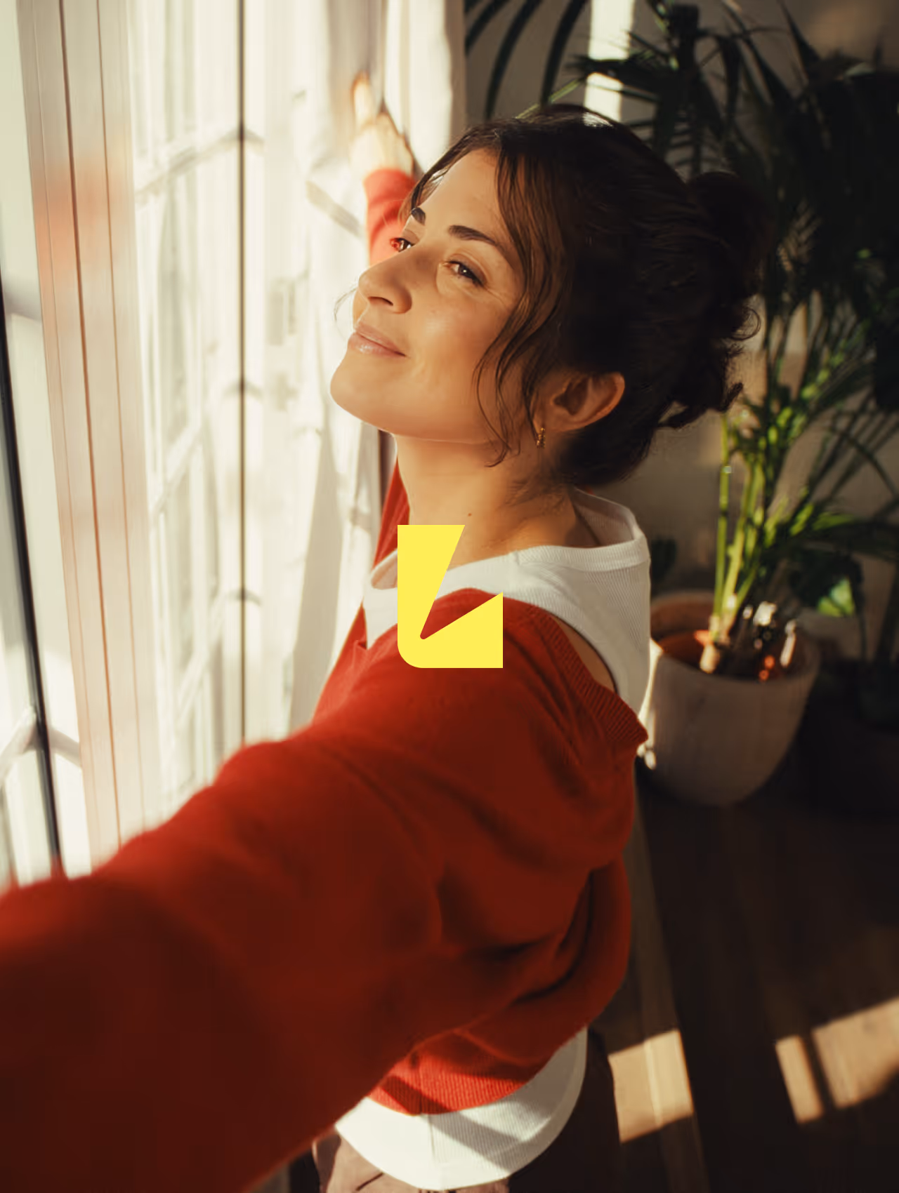 Woman wearing a red sweater stretching with arms wide open near a sunlit window and smiling.