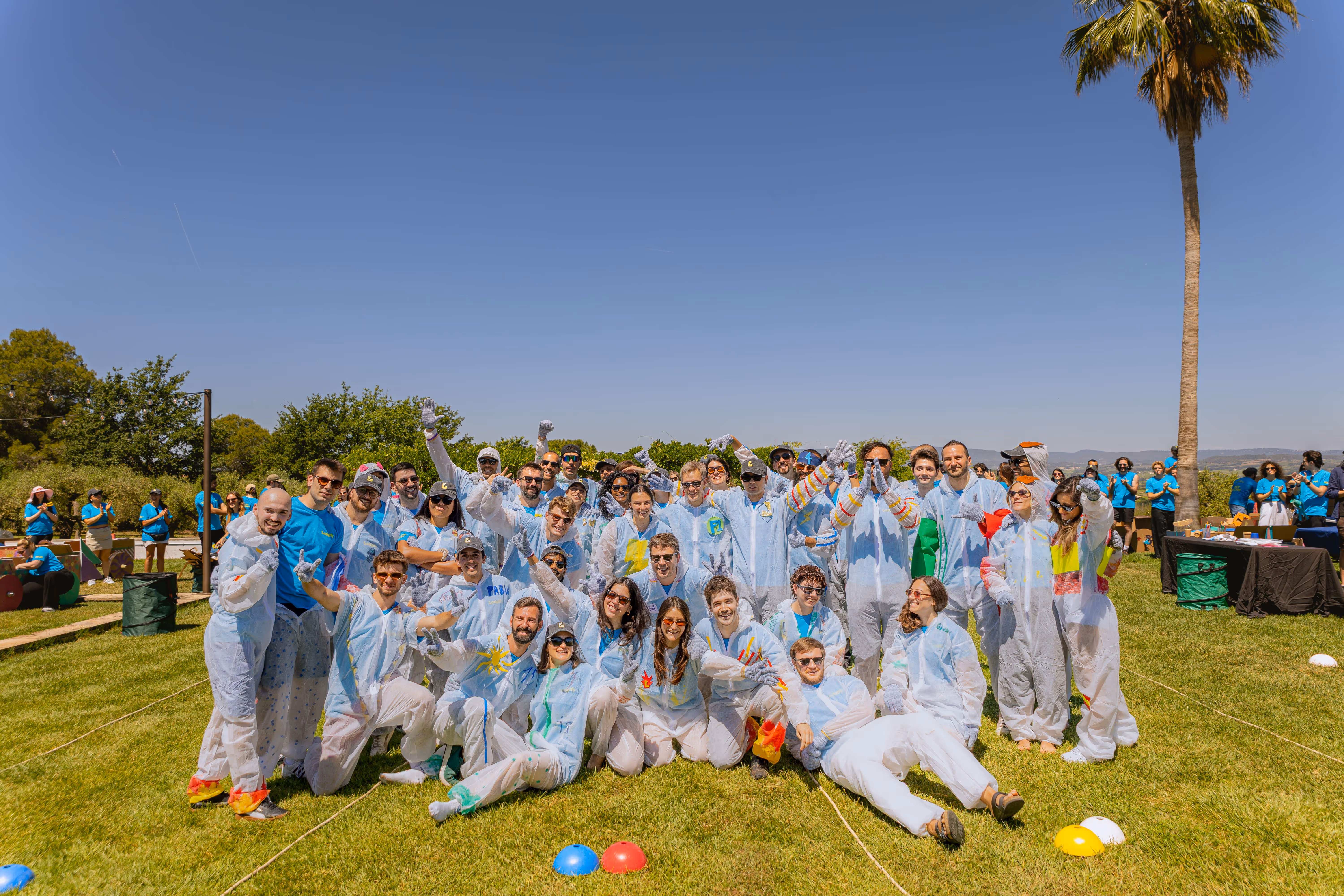 Group of people in white coveralls posing on grass on a sunny day with trees and clear blue sky.