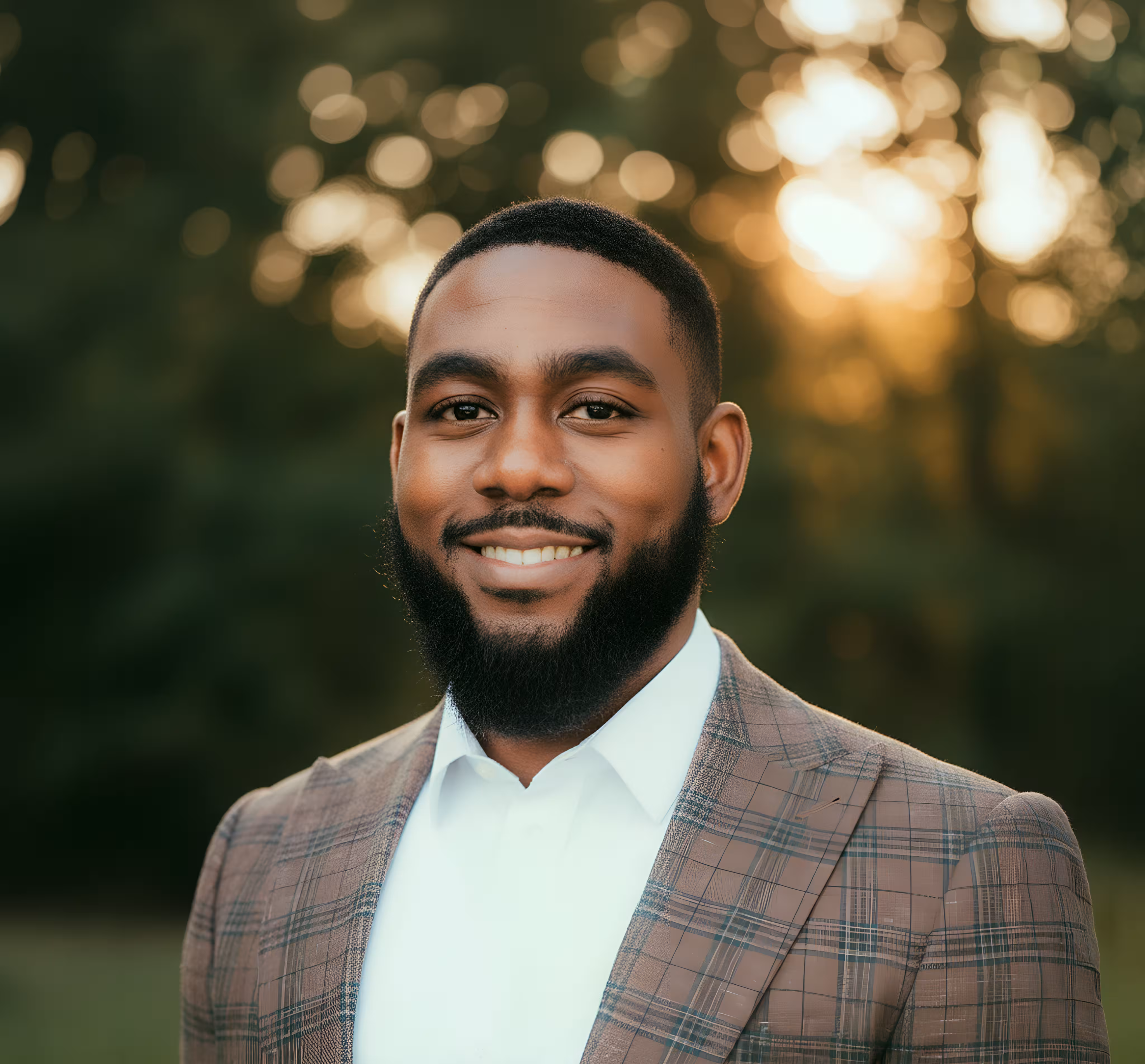 Smiling bearded man in a brown plaid blazer and white shirt outdoors with blurred greenery background.