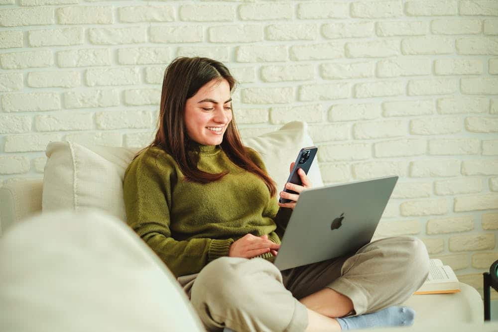 Smiling woman wearing green sweater using phone and laptop while sitting on couch against white brick wall.