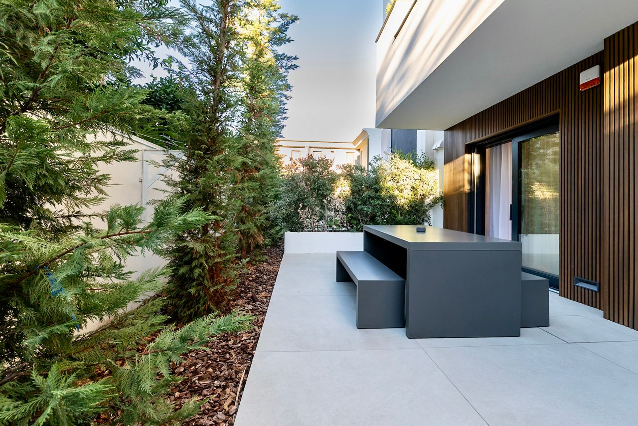 Modern patio with gray table and benches beside a house with wood paneling and greenery.