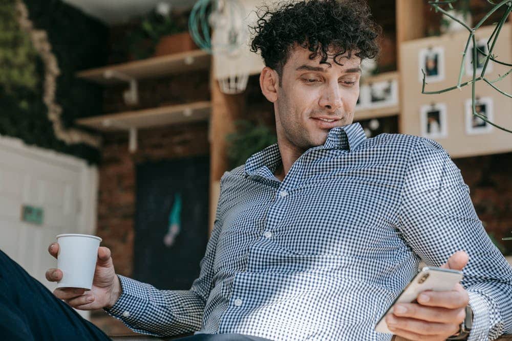 Man with curly hair in a checkered shirt holding a smartphone and a white cup indoors.