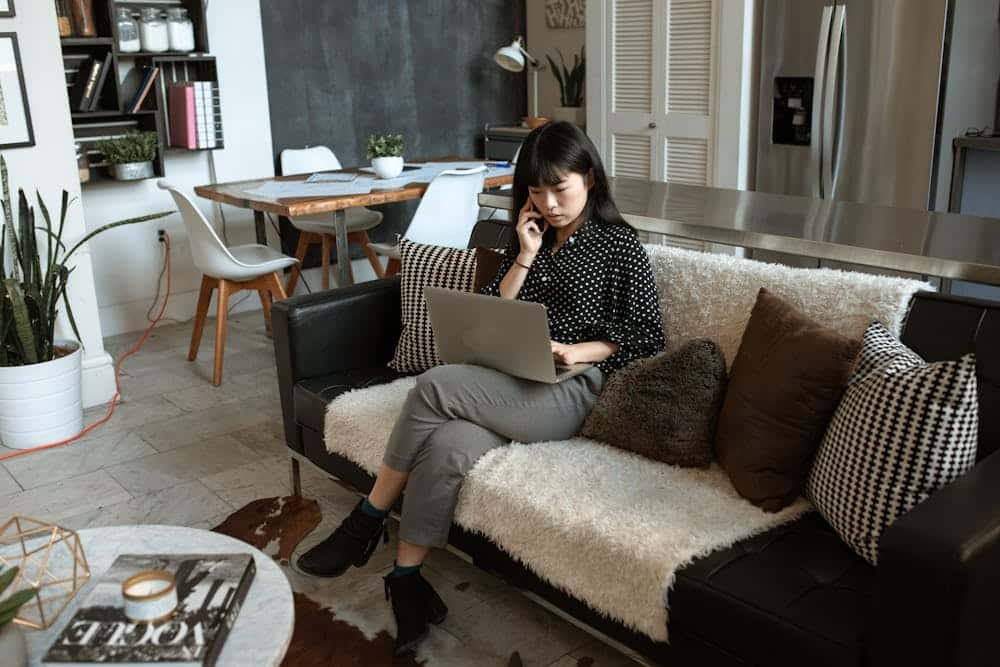 Woman sitting on a sofa using a laptop and talking on the phone in a modern living room.