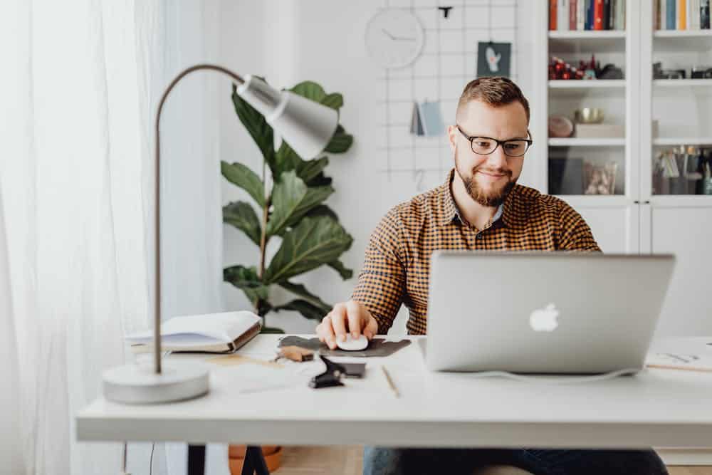 Man with glasses using a MacBook laptop at a cluttered desk with lamp and leafy plant.