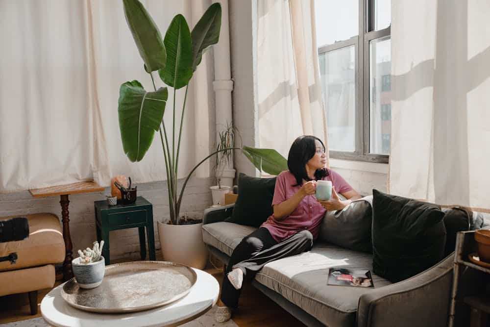 Woman in purple shirt sitting on gray couch holding a mug, looking out sunlit window with plants nearby.
