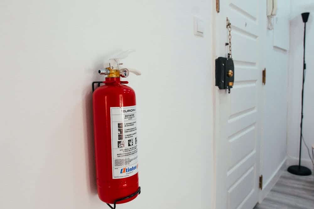 Red fire extinguisher mounted on a white wall next to a white door and black floor lamp.