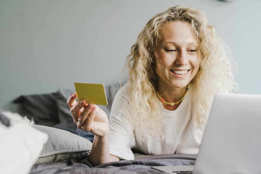 Smiling woman with curly hair holding a credit card and looking at a laptop on bed.