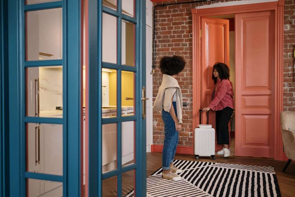 Two women with curly hair in a room, one holds a suitcase near an open coral door.
