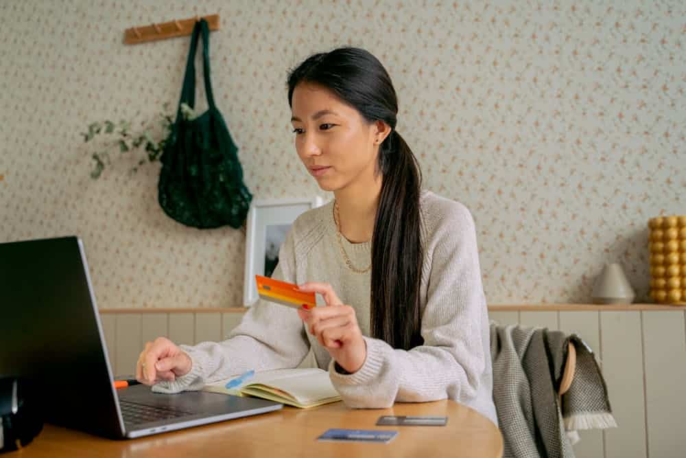 Woman holding a credit card and using a laptop at a wooden table with notebook and cards.
