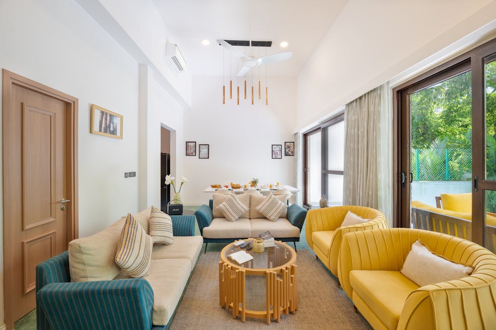 Bright living room with beige and yellow sofas, glass coffee table, and large windows with garden view.