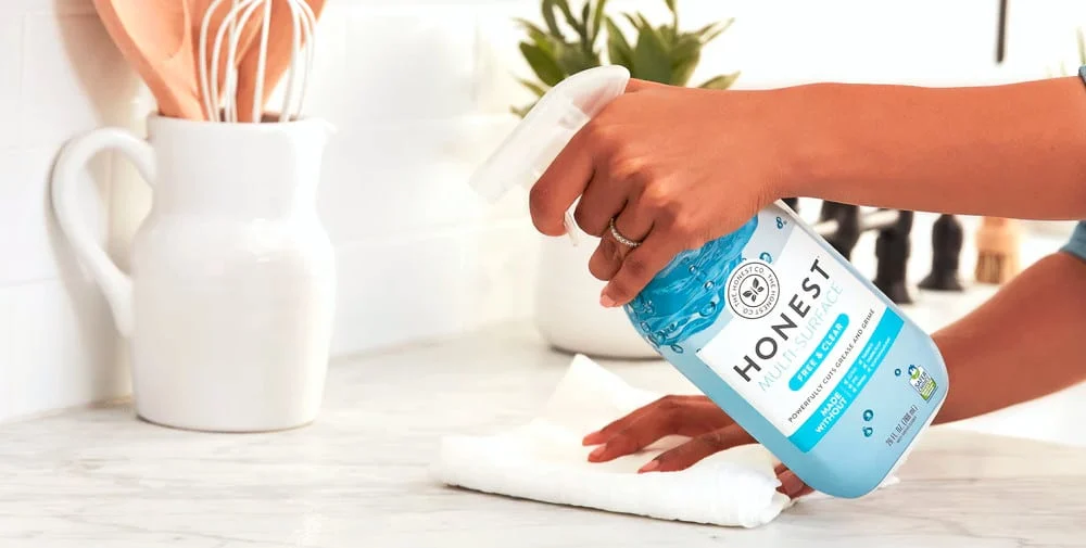 Person spraying Honest Multisurface Cleaner on white cloth to clean a light-colored countertop.