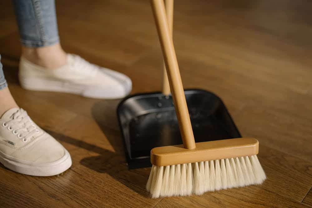 Person sweeping wooden floor with broom and dustpan wearing white sneakers and blue jeans.