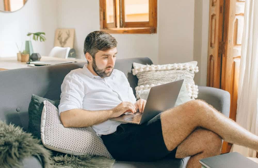 Man in white shirt and black shorts sitting on gray couch using a laptop in a bright room.