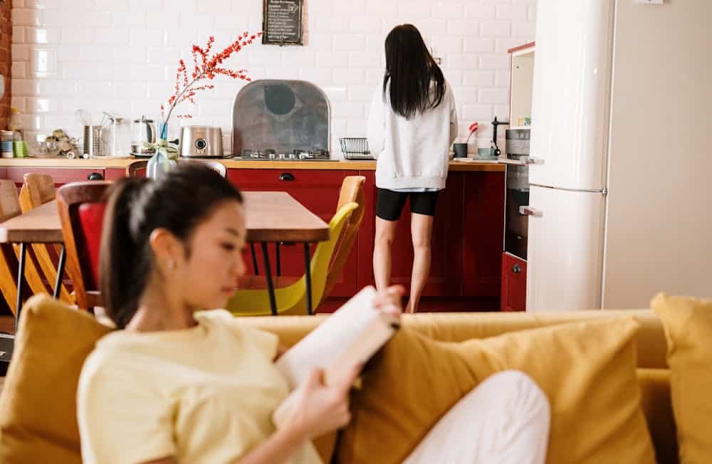 Woman reading on a yellow couch in the foreground, another standing in a red kitchen in the background.