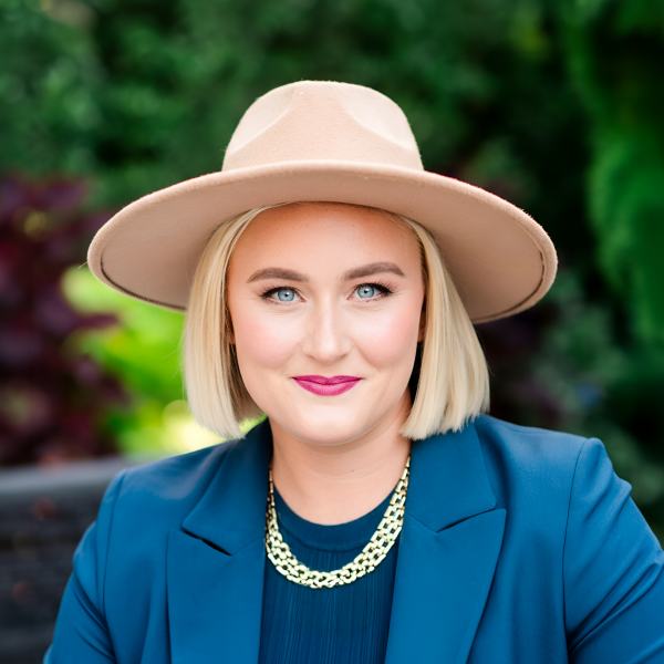 Blonde woman with blue eyes wearing a beige hat, blue blazer, and gold necklace outdoors.