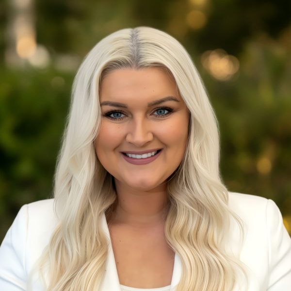Smiling woman with long wavy blonde hair wearing a white blazer, blurred greenery background.