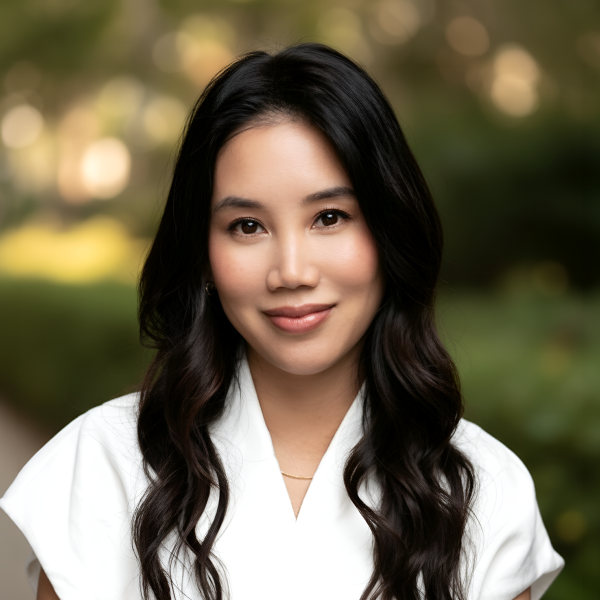 Portrait of a smiling woman with long dark hair in a white top, blurred natural background.