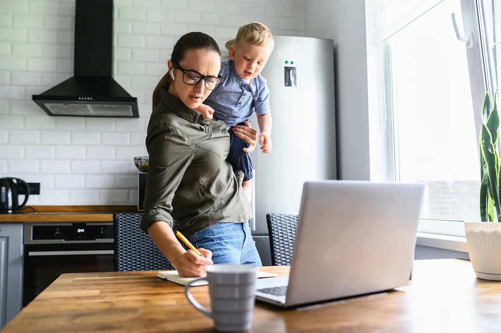 A busy mother balancing childcare and remote work in a kitchen environment.