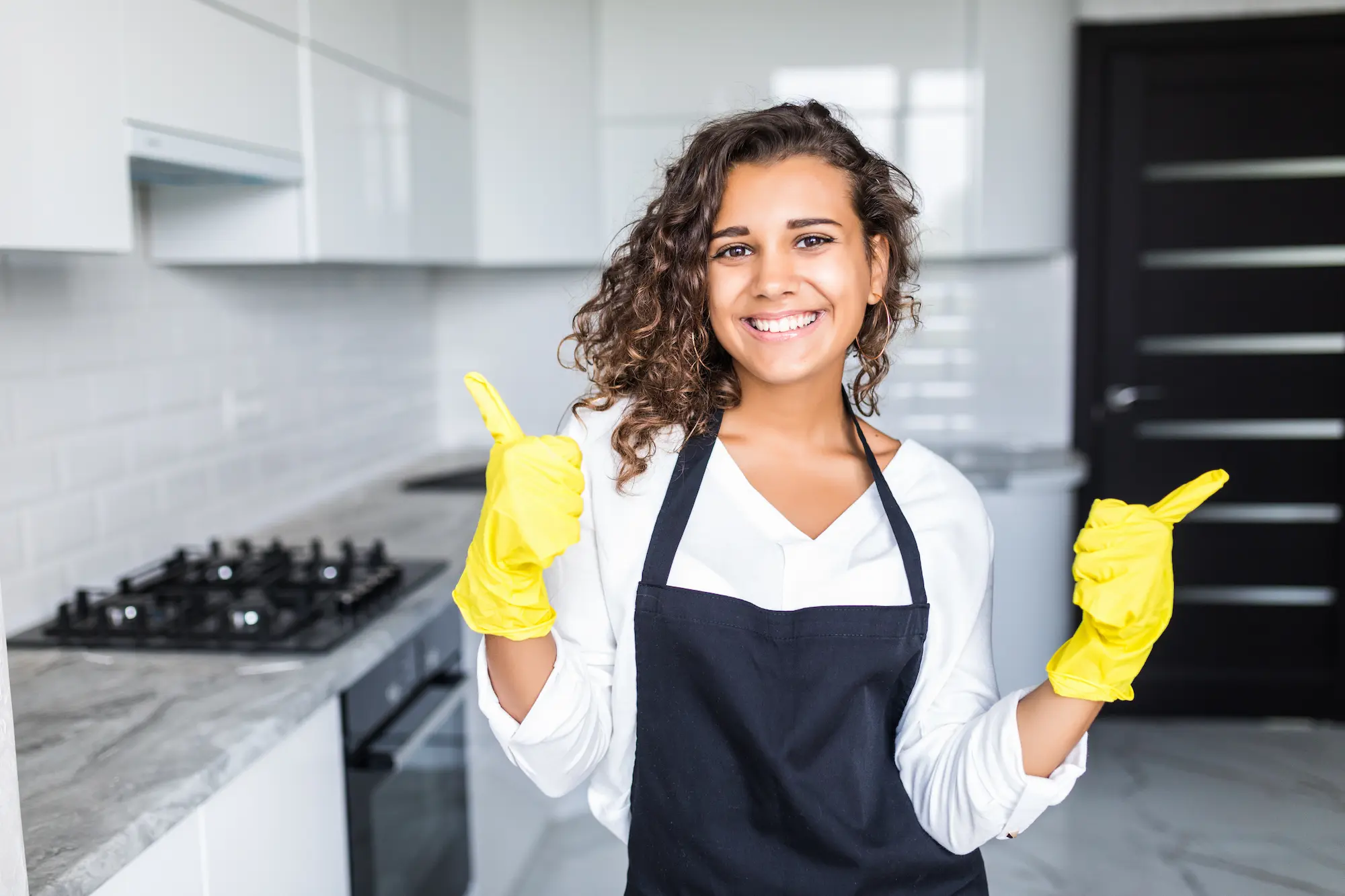 A smiling professional home cleaner wearing a black apron and yellow cleaning gloves.