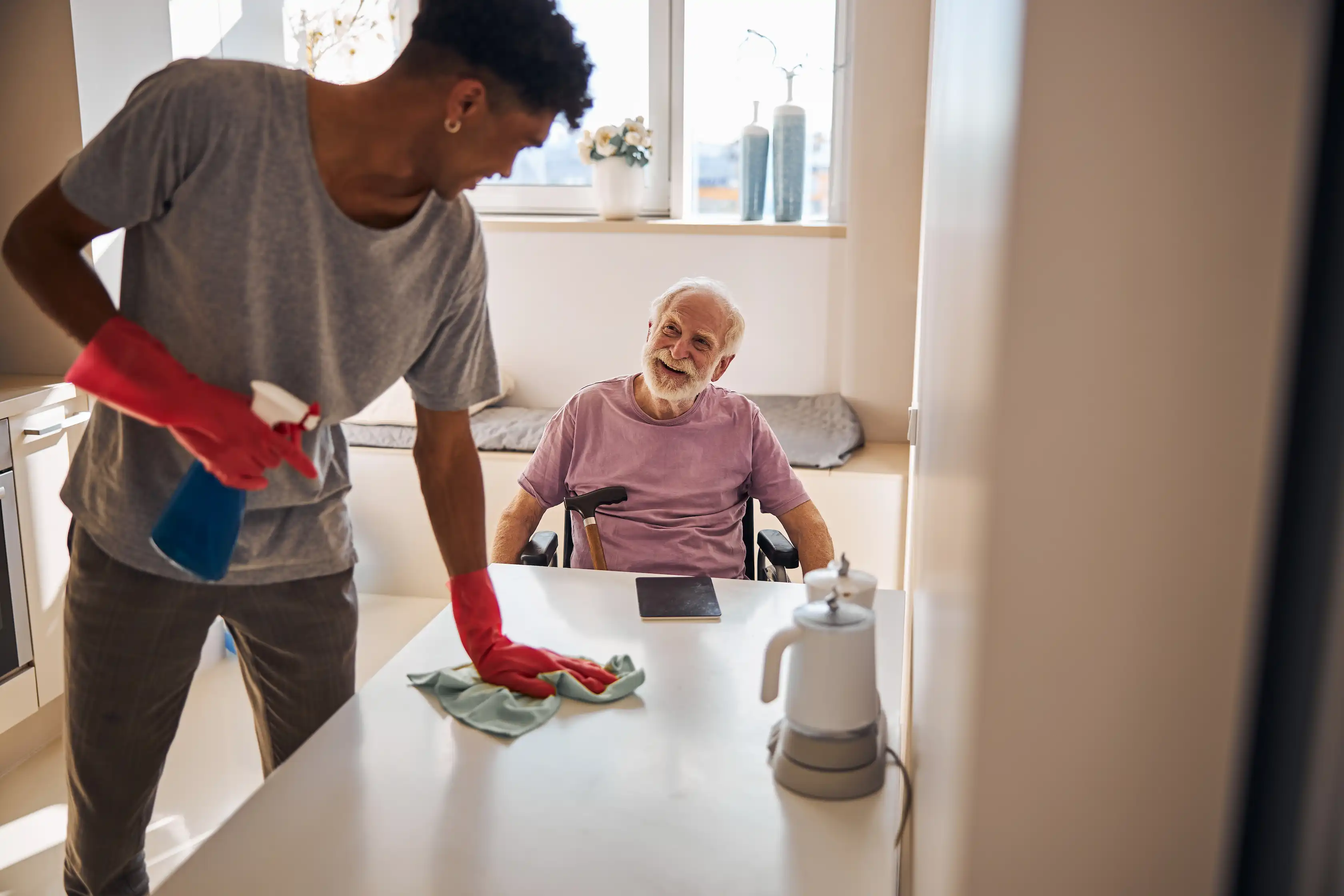 A support worker cleaning a table surface while interacting with an elderly man in a wheelchair.